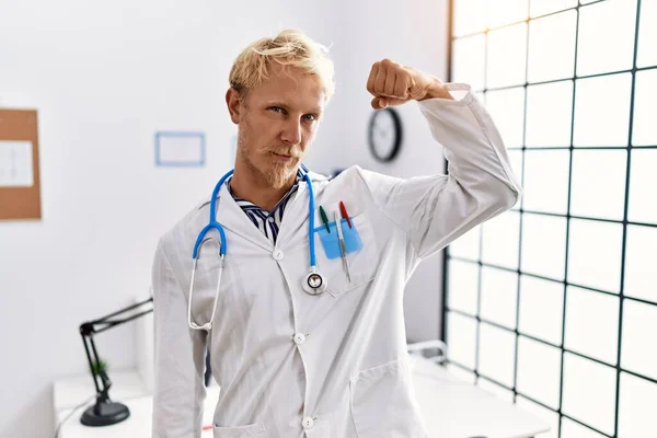 年轻金发男子身穿医生制服和听诊器在诊所强壮的人展示手臂肌肉 自信和自豪的力量 — 图库照片