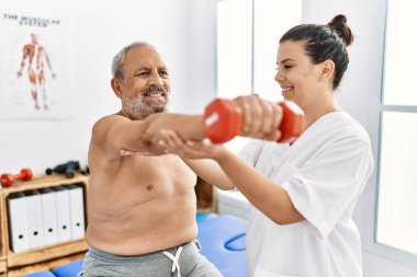 Fizyoterapist ve hasta gülümsüyor. Klinikteki halteri kullanarak rehabilitasyon seansına katılıyorlar.