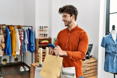 Genç İspanyol müşteri gülümsüyor alışveriş torbaları tutuyor ve giyim mağazasında akıllı telefon kullanıyor..