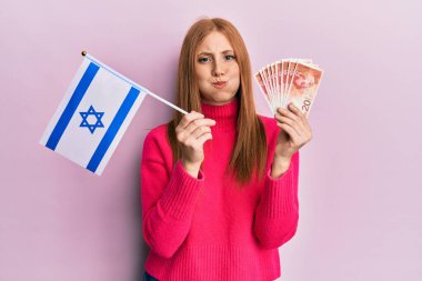 Genç İrlandalı kadın İsrail bayrağı ve şekel banknotları tutuyor komik suratıyla yanaklarını şişiriyor. Ağzı havayla şişer, havayı yakalar.. 