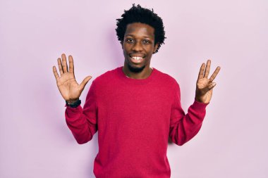 Genç, Afro-Amerikalı, günlük giysiler giymiş, 8 numaralı parmaklarıyla işaret ederken, kendinden emin ve mutlu bir şekilde gülümsüyor.. 