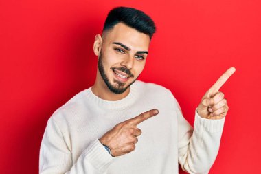 Sakallı, sıradan bir süveter giyen, gülümseyen ve kameraya bakan, iki eli ve parmağı yanda olan genç İspanyol bir adam.. 