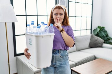 Genç kızıl saçlı kadın elinde plastik şişelerle çöp sepetini tutarak parmakları dudaklarında sessiz olmak istiyor. sessizlik ve gizli konsept. 