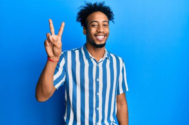 Sakallı, sıradan çizgili tişörtlü, genç, Afro-Amerikan bir adam. İki numaralı parmağını gösterip kendini beğenmiş ve mutlu bir şekilde gülümsüyor.. 