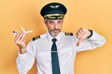 Yakışıklı, orta yaşlı, gri saçlı, uçak pilotu üniforması giyen, kızgın yüzlü oyuncak uçak tutan, olumsuz işaretli, baş parmakları aşağıda, reddedilme konsepti olan bir adam. 