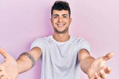 Latin asıllı genç bir adam, günlük beyaz tişört giyip kameraya bakıyor ve sarılmak için kollarını açmış gülümsüyor. Mutluluğu kucaklayan neşeli bir ifade.. 
