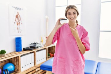 Ağrı kesici kliniğinde çalışan genç fizyoterapist kadın elinde kredi kartıyla mutlu mesut elini ve parmağını göstererek gülümsüyor. 