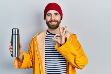 Beyaz, sakallı, termosu olan bir adam parmaklarıyla imza atıyor, gülümseyerek mükemmel bir sembol oluşturuyor. 