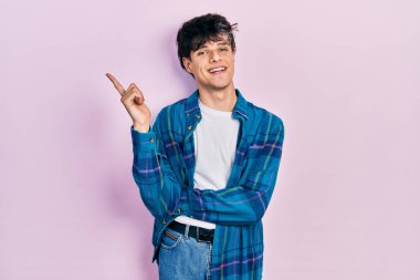 Yakışıklı hippi genç adam, günlük beyaz tişört ve klasik gömlek giyiyor. Yüzünde kocaman bir gülümseme var. Eli ve parmağı kameraya bakıyor.. 