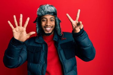Sakallı, kışlık şapkalı ve paltolu Afro-Amerikalı genç adam yedinci parmaklarını gösterirken kendinden emin ve mutlu bir şekilde gülümsüyor.. 