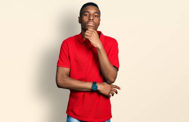 Genç Afro-Amerikalı bir adam, elleri çenesinde gündelik kırmızı tişört giyip soru sormayı düşünüyor, düşünceli bir ifade. Düşünceli bir yüzle gülümsüyor. şüphe kavramı. 