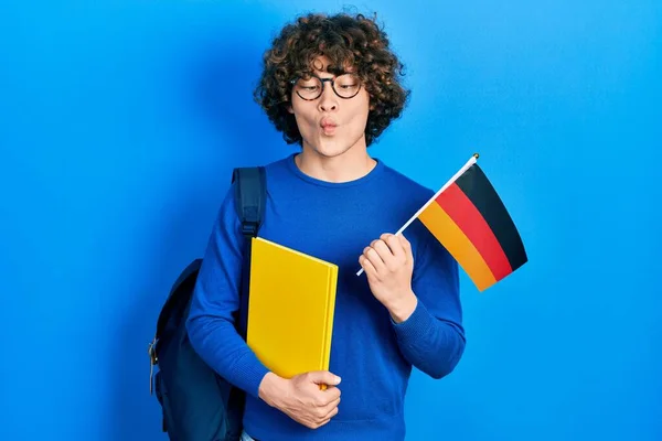 Handsome young man exchange student holding germany flag making fish face with mouth and squinting eyes, crazy and comical.