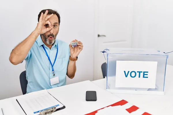 一个留着胡子的中年男子举着选票坐着我的投票徽章做着一个很好的手势 脸上带着惊讶的表情 眼睛透过手指看着 不信的表情 — 图库照片