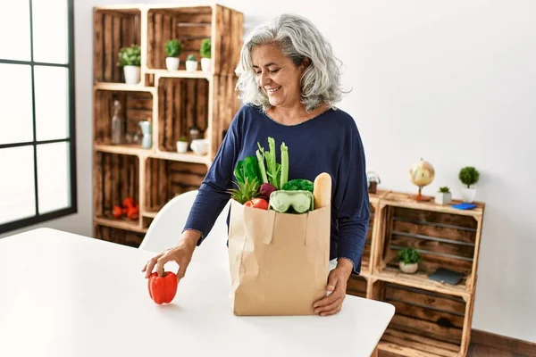Middle age grey-haired woman holding paper bag with groceries standing at home.