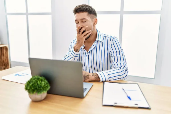 年轻英俊的男人 留着胡子 在办公室里用电脑笔记本电脑工作 厌烦了打呵欠 累了用手捂住嘴 烦躁不安和困倦 — 图库照片