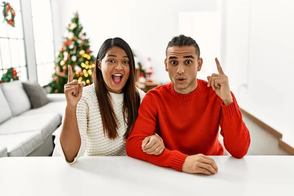 Young Latin Couple Sitting Table Christmas Tree Pointing Finger Successful — Zdjęcie stockowe