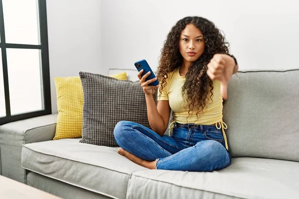 スマートフォンを使って自宅のソファに座っている美しいヒスパニック系の女性は 親指を下にジェスチャーで不満と否定を示して怒っているように見えます 悪い表現 — ストック写真