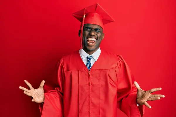 卒業キャップと儀式用のローブを着た若いアフリカ系アメリカ人男性が腕を上げて興奮して叫んで目を閉じて成功のために狂気と狂気を祝う 勝者のコンセプト — ストック写真