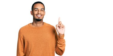 Genç Afro-Amerikalı günlük kıyafetler giymiş bir numaralı parmağını gösterip işaret ederken kendinden emin ve mutlu bir şekilde gülümsüyor.. 