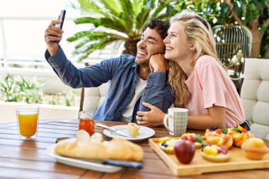 Genç çift kahvaltı yapıyor ve akıllı telefon başında özçekim yapıyor terasta masada oturuyor..