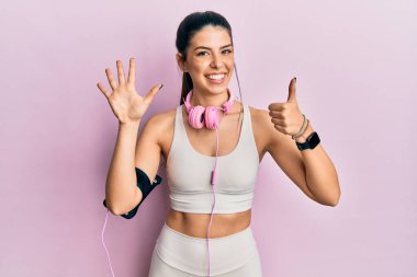 Spor kıyafetleri giyen ve kulaklık takan İspanyol genç bir kadın altı numaralı parmaklarını gösterip işaret ederken kendinden emin ve mutlu bir şekilde gülümsüyor.. 