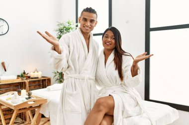 Young latin couple wearing towel standing at beauty center smiling cheerful presenting and pointing with palm of hand looking at the camera. 