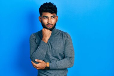 Balıkçı yaka kazak giyen, eli çenesinde soru soran Arap bir adam. Düşünceli bir ifade. Gülümseyen ve düşünceli bir yüz. şüphe kavramı. 