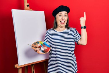 Orta yaşlı İspanyol kadın paletle resim çiziyor, resim sehpası ayakta şaşkın şaşkın gülüyor, parmaklarıyla işaret ediyor ve kollarını kaldırıyor.. 