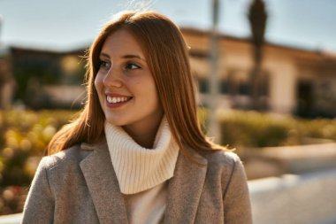 Genç kızıl kız gülümsüyor, mutlu bir şekilde şehirde duruyor..