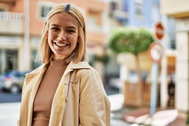 Genç sarışın kız gülümsüyor, mutlu bir şekilde şehirde duruyor..