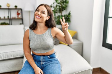 Genç esmer kadın evdeki kanepede oturmuş gülümsüyor ve parmaklarını zafer işareti yaparken kameraya bakıyor. İki numara.. 