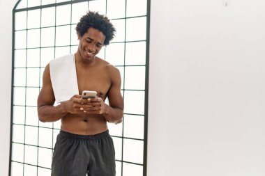 Genç Afrikalı Amerikalı adam spor salonunda akıllı telefon kullanarak mutlu bir şekilde gülümsüyor.