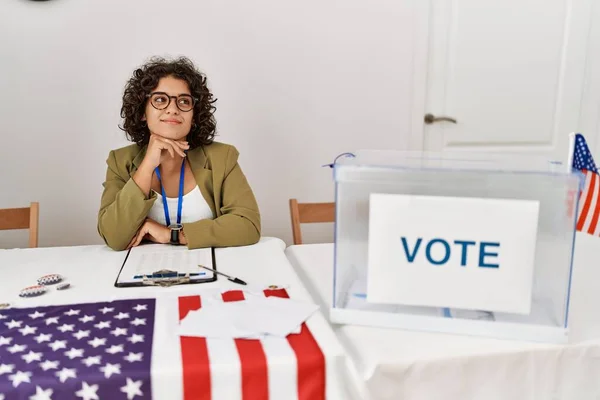 Young hispanic woman at political election sitting by ballot with hand on chin thinking about question, pensive expression. smiling with thoughtful face. doubt concept.