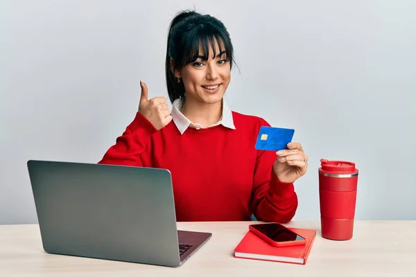 Young brunette woman with bangs working at the office using laptop and credit card smiling happy and positive, thumb up doing excellent and approval sign