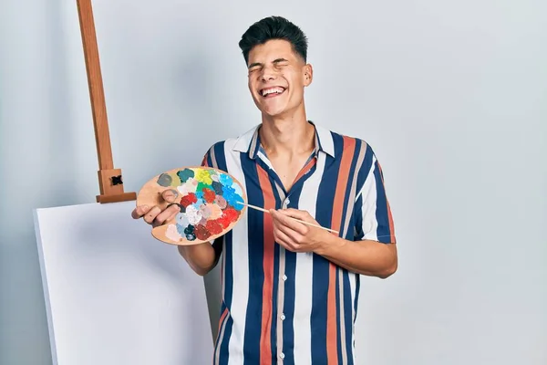 Jovem Hispânico Segurando Paleta Pintor Pincel Perto Tela Sorrindo Rindo — Fotografia de Stock