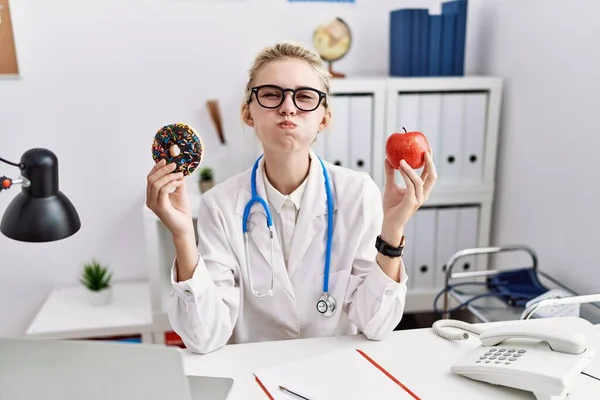年轻的女医生拿着红苹果和甜甜圈站在诊所里 用滑稽的脸鼓起双颊 嘴被空气吹胀 — 图库照片