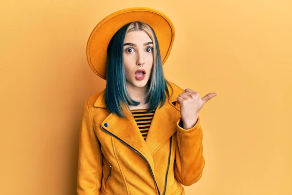 若い現代の女の子を身に着けている黄色の帽子と革のジャケット驚きのポインティングで手指で側に 口を開けて驚きの式 — ストック写真
