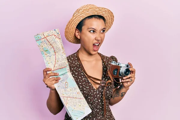 Beautiful hispanic woman with short hair holding city map and vintage camera angry and mad screaming frustrated and furious, shouting with anger. rage and aggressive concept.