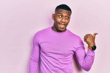 Genç siyah bir adam, günlük pembe süveter giyiyor, gülümsüyor, gülümsüyor ve başparmağıyla yan tarafa bakıyor.. 