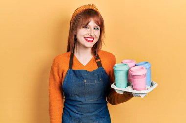 Kızıl saçlı, garson önlüğü giymiş genç bir kadın elinde kahve fincanları taşırken pozitif ve mutlu görünüyor ve gülümsüyor dişlerini gösteren bir gülümsemeyle. 