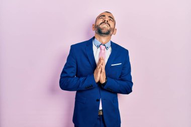 Genç İspanyol bir adam iş elbisesi ve kravat takıyor. Elleriyle yalvarıyor ve dua ediyor. Yüzünde umut ifadesi var. Yalvarıyor. 