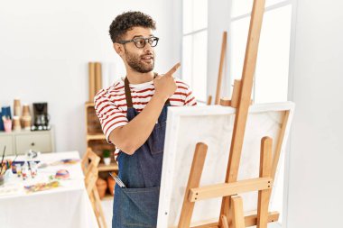 Sanat stüdyosundaki Arap genç adam yüzünde kocaman bir gülümsemeyle, kameraya el ile işaret ederek bakıyor.. 