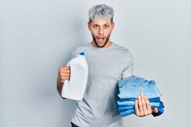 Çamaşır ve deterjan şişesi için kot pantolon tutan İspanyol asıllı genç adam şaşkınlık ve şaşkınlık ifadesi, korku ve heyecanlı yüz ifadesiyle şok oldu ve korktu.. 