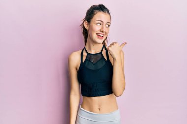 Genç esmer kadın spor giysisi giymiş mutlu yüzüyle gülümseyen ve başparmağıyla yan tarafı gösteren.. 