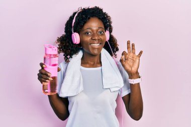Genç Afro-Amerikalı kadın spor kıyafeti giymiş elinde su şişesi kulaklık takıyor parmaklarıyla imza atıyor, gülümseyerek mükemmel bir sembol oluşturuyor. 