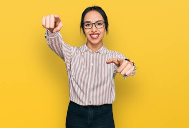 Gündelik kıyafetler giyen ve gözlüklü genç İspanyol kız seni ve kamerayı işaret ediyor, gülümsüyor, pozitif ve neşeli. 