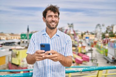 Genç İspanyol adam eğlence parkında akıllı telefon kullanarak mutlu bir şekilde gülümsüyor..