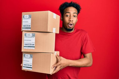 Sakallı, elinde teslimat paketleri olan afro-amerikalı genç bir adam şaşkınlık ve şaşkınlık dolu bir ifade, korku ve heyecanlı bir yüz ifadesiyle. 