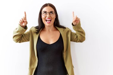 Genç esmer kadın izole edilmiş arka planda duruyor. Şaşırmış, şaşırmış, parmaklarıyla işaret ediyor ve kollarını kaldırıyor.. 