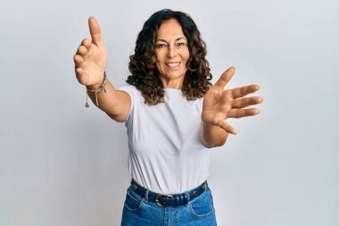Orta yaşlı İspanyol bir kadın, günlük beyaz tişört giyiyor kameraya bakıyor ve sarılmak için kollarını açmış gülümsüyor. Mutluluğu kucaklayan neşeli bir ifade.. 